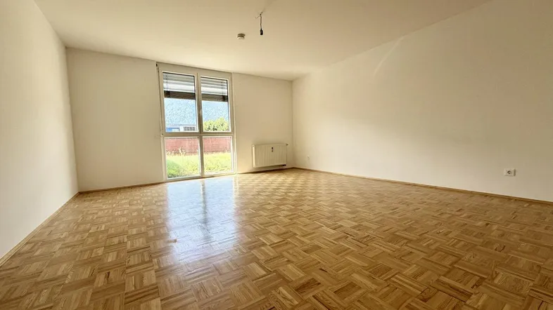 Helles, leeres Wohnzimmer mit Parkettboden und Zugang zu einem Außenbereich.
