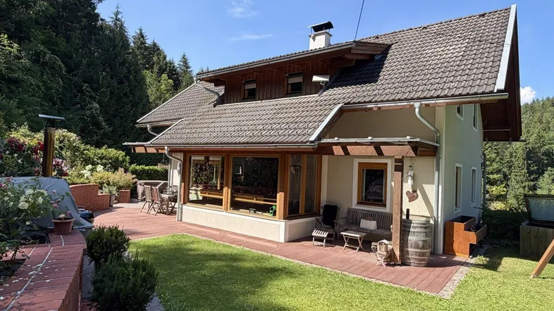 Haus mit großer gepflasterter Terrasse, Rasenfläche und umgeben von dichtem Wald, ideal für Entspannung im Freien.