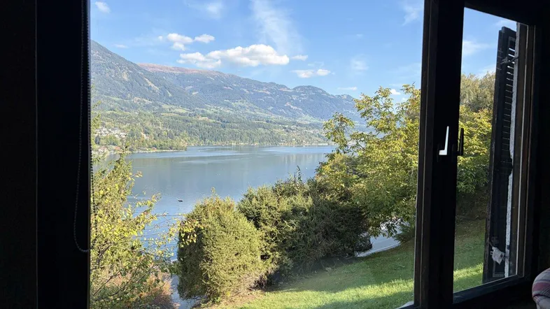 Panoramablick auf den See und die Berge durch ein großes Fenster, umgeben von üppigem Grün.