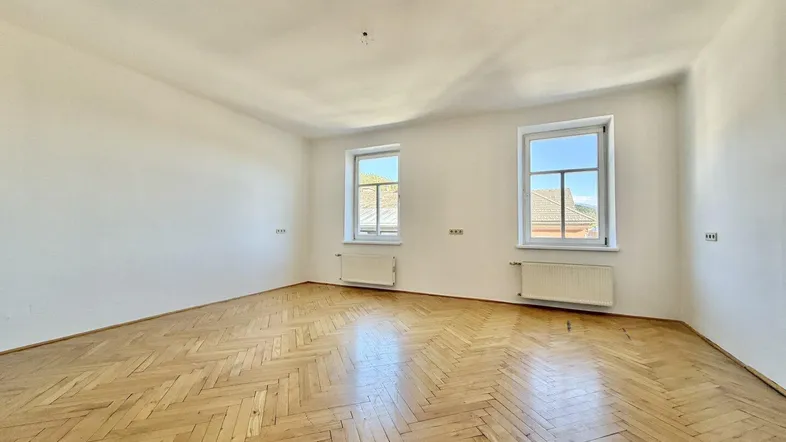 Geräumiger Raum mit Fischgrätparkettboden, weißen Wänden und zwei Fenstern, die viel Tageslicht hereinlassen.