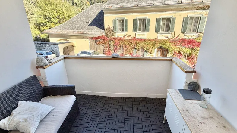 Kleiner, möblierter Balkon mit Blick auf ein charmantes Gebäude und grüne Hügel.