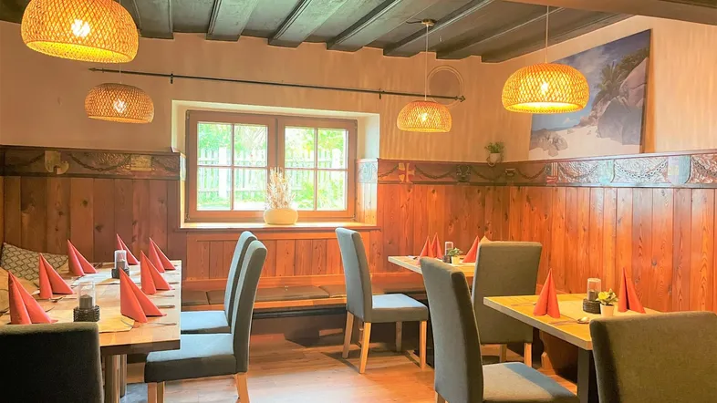 Gemütlicher Essbereich mit Holztäfelung, Holzbalkendecke und geflochtenen Pendelleuchten, durch ein Fenster fällt Tageslicht herein.