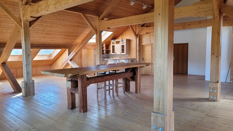 Geräumiger offener Wohnbereich mit sichtbaren Holzbalken, Holzfußboden und einem großen Holztisch unter Dachfenstern.