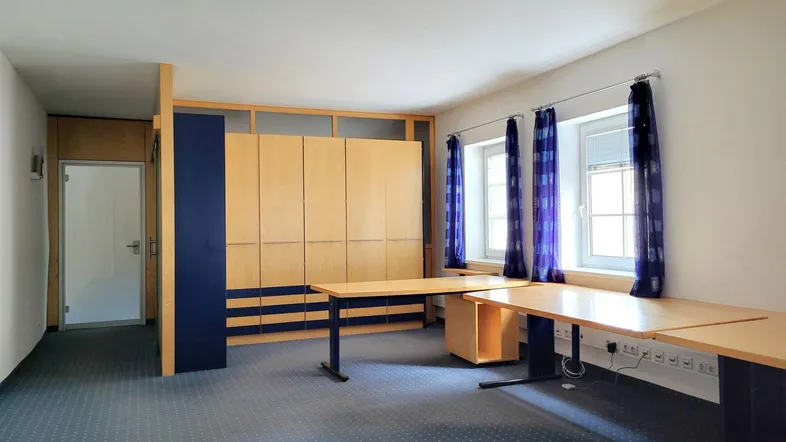 Geräumiges Büro mit blaugrauem Teppichboden, Holzmöbeln und großen Fenstern mit Vorhängen.