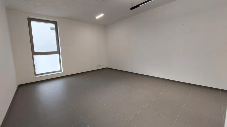 Leerer Büroraum mit großen Fenstern, grauen Fliesen und moderner Deckenbeleuchtung, ideal für gewerbliche Nutzung.