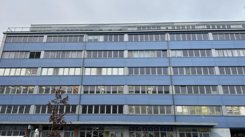 Blaues Geschäftsgebäude mit vielen Fenstern und Autos auf der Straße davor, unter bewölktem Himmel.