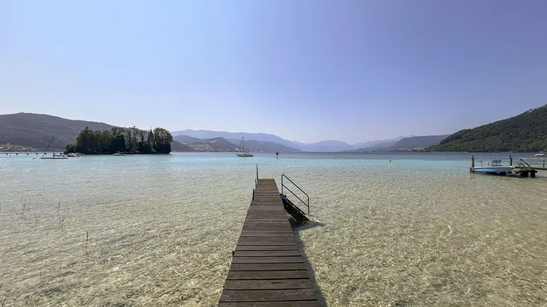 Langer Holzsteg führt in einen klaren See mit malerischer Bergkulisse und kleiner Insel.