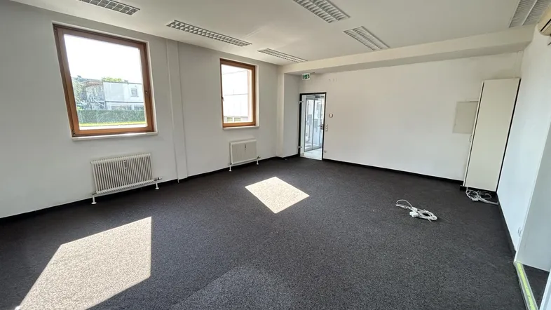 Geräumiges, leeres Büro mit Teppichboden, großen Fenstern und Heizkörpern, gut beleuchtet.