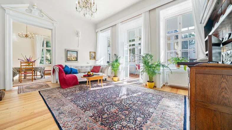 Helles Wohnzimmer mit Holzboden, großem Teppich, Kronleuchter und Zugang zu einem weiteren Raum.