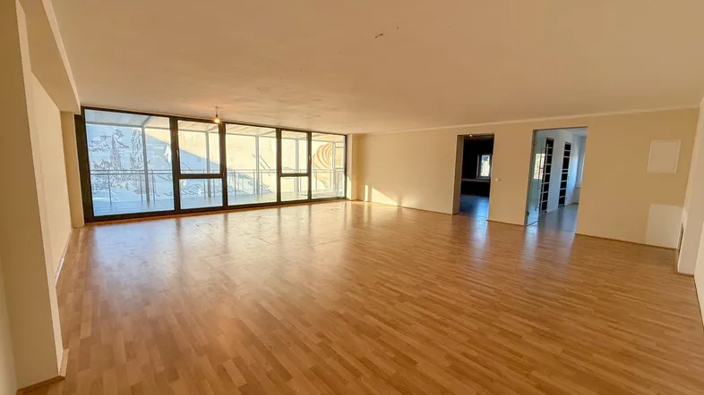 Geräumiger, leerer Wohnbereich mit großen Fenstern und Holzboden, der viel Tageslicht hereinlässt.