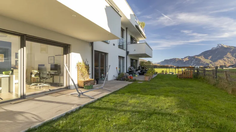 Moderne Wohnanlage mit Terrasse, Rasenfläche und Bergblick unter strahlend blauem Himmel.