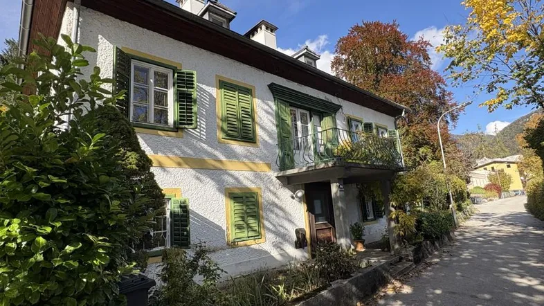 Traditionelles Haus mit weißer Fassade, grünen Fensterläden und einem kleinen Balkon unter blauem Himmel.