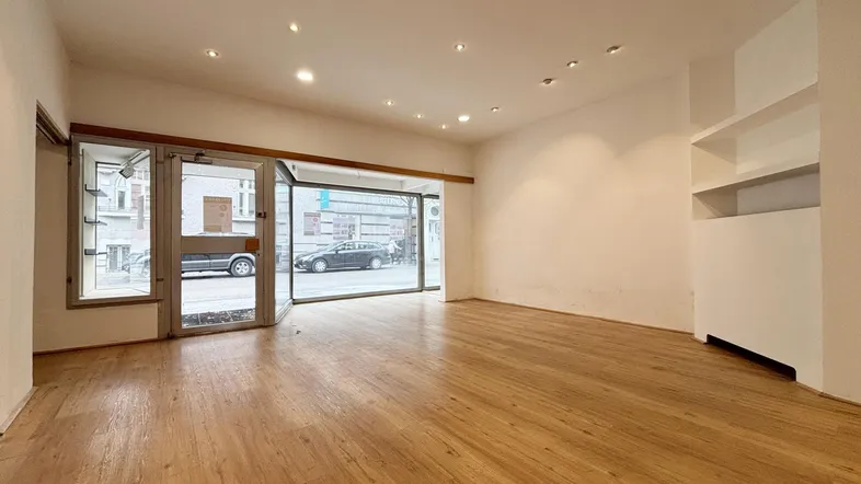 Heller, leerer Ladenraum im Erdgeschoss mit großen Schaufenstern zur Straße und Holzboden.
