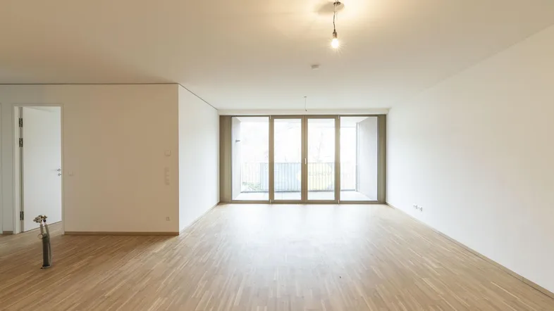 Heller, leerer Wohnbereich mit Parkettboden und großen Schiebetüren zum Balkon.