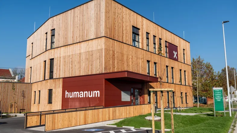 Modernes Gebäude mit Holzfassade und barrierefreiem Zugang, umgeben von Grünflächen unter blauem Himmel.