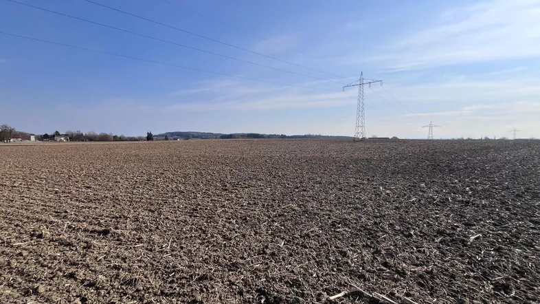 Weites Feld unter blauem Himmel mit Stromleitungen und einem Sendemast in der Ferne.