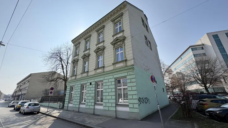 Historisches Gebäude mit hellgrüner und beiger Fassade an einer Straße mit geparkten Autos unter blauem Himmel.