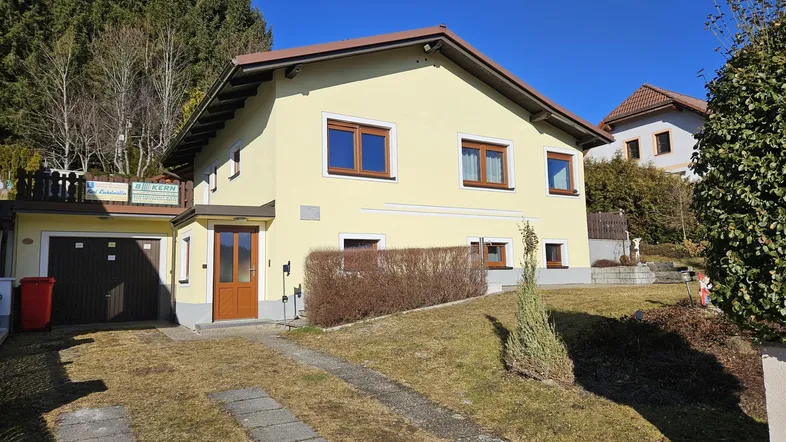 Gelbes Einfamilienhaus mit braunen Fenstern, Garage und Auffahrt, umgeben von Bäumen.