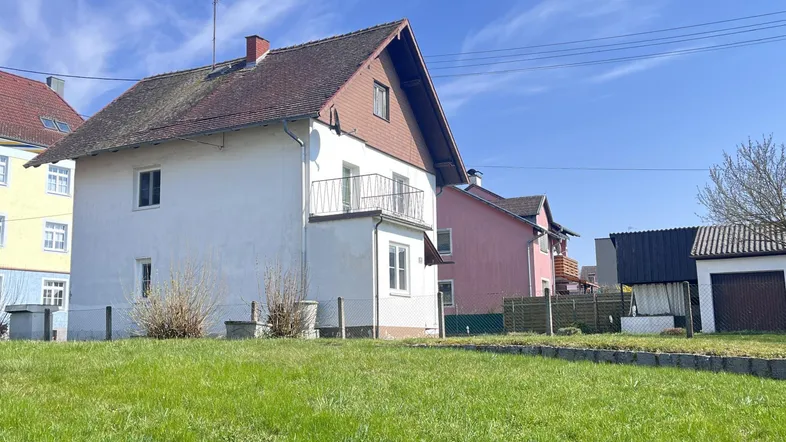 Ansicht des Wohnhauses von außen mit weißer Fassade, Balkon und Satteldach unter blauem Himmel.
