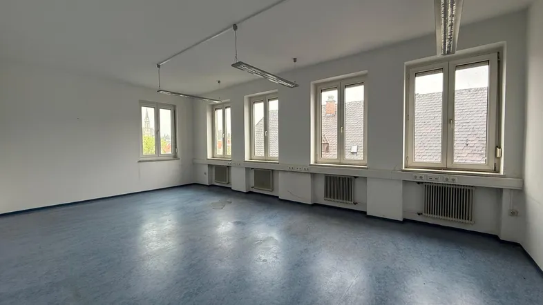 Geräumiger, leerer Büroraum mit blauer Bodenfläche, weißen Wänden und mehreren Fenstern für Tageslicht.