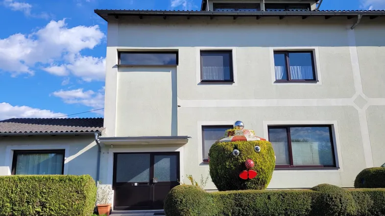 Hausfassade mit hellen Wänden, dunklen Fenstern und gepflegter Hecke im Vorgarten unter blauem Himmel.