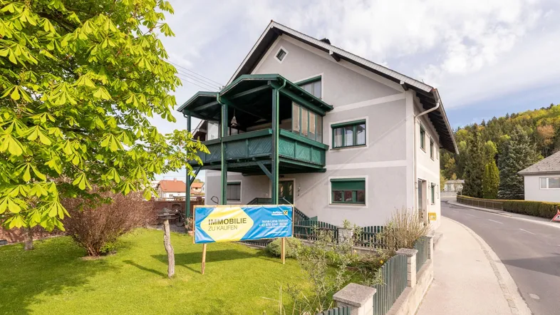 Graues Haus mit grünem Balkon und Vorgarten, umgeben von Bäumen und einer Straße.