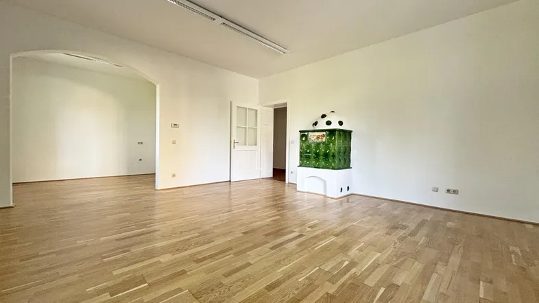 Geräumiges Zimmer mit hellem Parkettboden, weißen Wänden und einem traditionellen grünen Kachelofen.