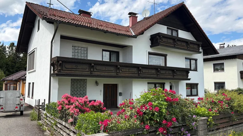Traditionelles Haus mit weißer Fassade, Holzbalkonen und blühenden Blumen im Vorgarten.