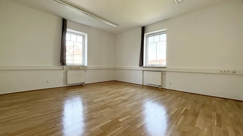 Helles, leeres Zimmer mit Parkettboden, weißen Wänden und zwei Fenstern, ideal als Büro.