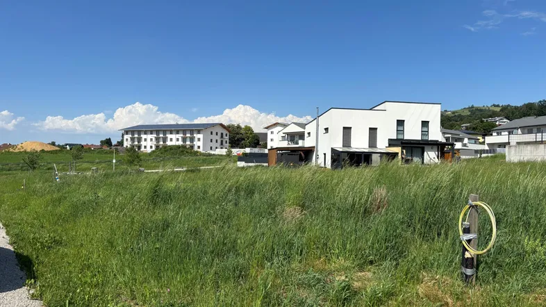 Weitläufiges grünes Feld mit modernen Gebäuden und Hügellandschaft im Hintergrund unter blauem Himmel.