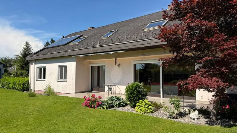 Hausfassade mit kleiner Terrasse, gepflegtem Rasen und Gartenbereich unter blauem Himmel.