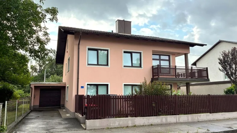 Einfamilienhaus mit rosafarbener Fassade, Balkon und integrierter Garage unter bewölktem Himmel.