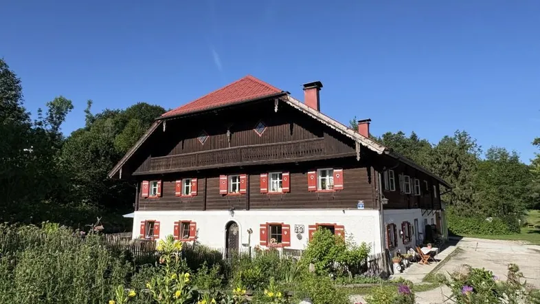 Traditionelles Bauernhaus mit rotem Dach, Holzfassade und gepflegtem Vorgarten unter blauem Himmel.