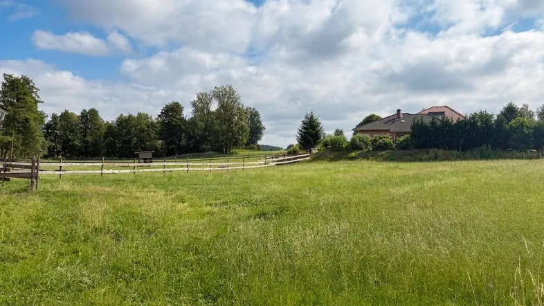 Weitläufige grüne Wiese mit einem Haus im Hintergrund und umzäunten Bereichen unter blauem Himmel.