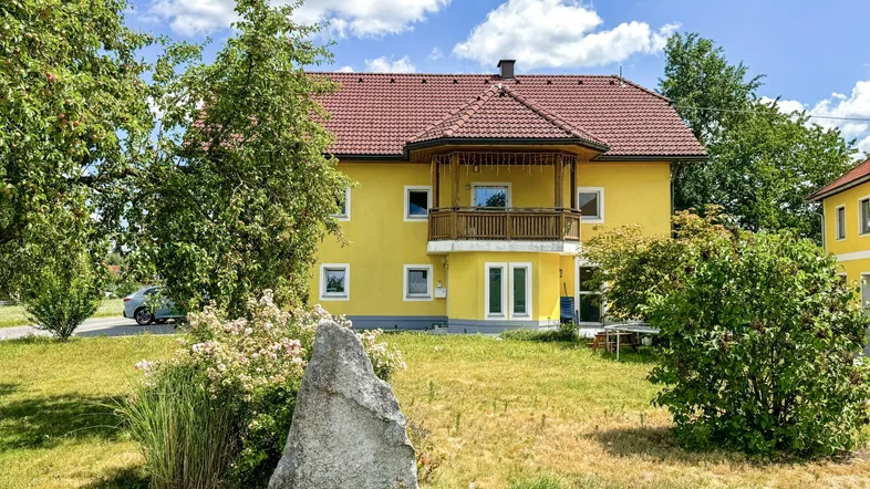 Gelbes Wohnhaus mit rotem Ziegeldach, Balkon und großem Vorgarten unter blauem Himmel.