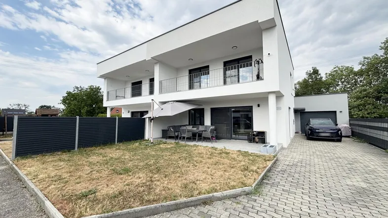 Moderne Doppelhaushälfte mit Terrasse, Garten und Auffahrt, umgeben von einer dunklen Zaunanlage.