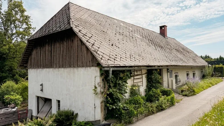 Ländliches Gebäude mit traditionellem Dach und bewachsenen Fassadenbereichen unter bewölktem Himmel.