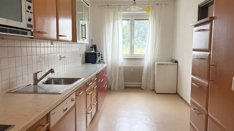 Funktionale Küche mit Holzschränken, weißer Arbeitsplatte, Spüle und Blick ins Grüne durch das Fenster.