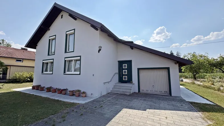 Weißes Einfamilienhaus mit dunkelgrünen Akzenten, gepflasterter Auffahrt und Garage unter strahlendem Himmel.