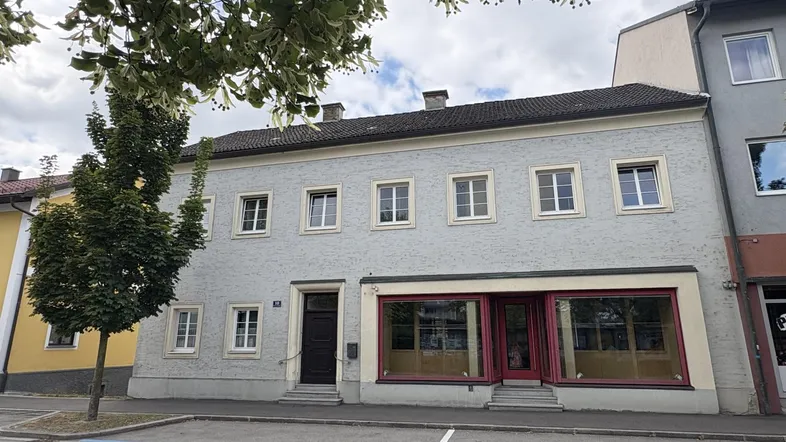 Vorderansicht eines älteren Gebäudes mit hellgrauer Fassade und großen Schaufenstern im Erdgeschoss.