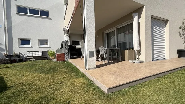 Große, geflieste Terrasse mit Gartenmöbeln und angrenzender Rasenfläche unter blauem Himmel.