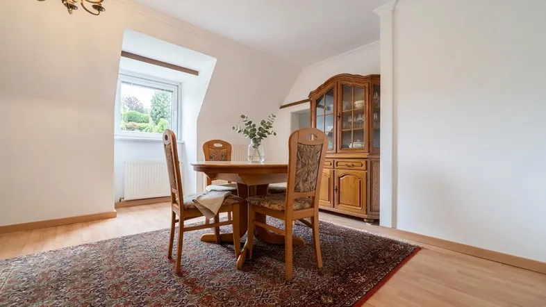 Esstisch mit Stühlen und einem traditionellen Holzschrank in einem hellen Raum mit Teppich.