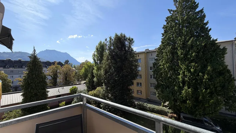 Sonniger Balkon mit Metallgeländer, bietet einen weiten Blick auf grüne Bäume und ferne Berge.
