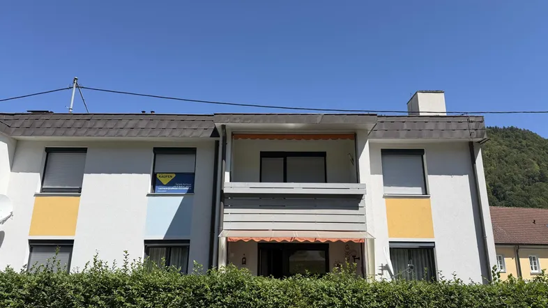 Mehrfamilienhaus mit weißer Fassade, farbigen Akzenten und Balkonen, umgeben von einer gepflegten Hecke und mit Bergblick.