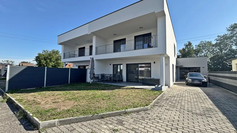 Modernes weißes Gebäude mit Flachdach, Balkonen, Terrasse und gepflasterter Auffahrt.