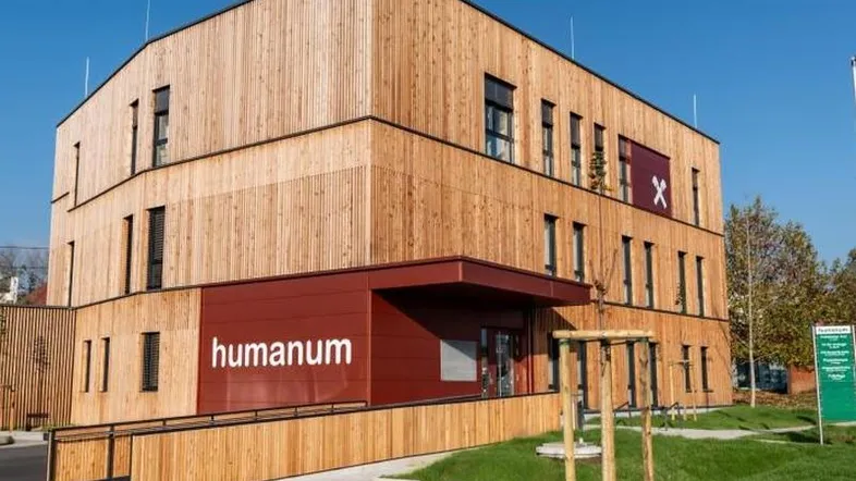 Modernes Gebäude mit Holzfassade und rotem Eingangsbereich, inklusive barrierefreier Rampe und Grünflächen.