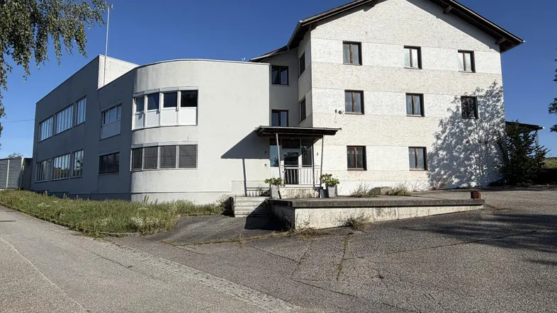Mehrteiliges Gebäude mit heller Fassade, großen Fenstern und einem geschwungenen Anbau in ländlicher Umgebung.