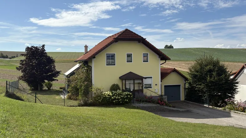 Gelbes Einfamilienhaus mit rotem Ziegeldach, umgeben von grünen Feldern unter blauem Himmel.
