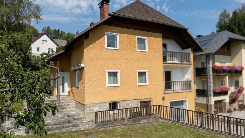 Gelb verputztes Haus mit braunem Dach, Balkonen und einem gepflegten Gartenbereich im Vordergrund.