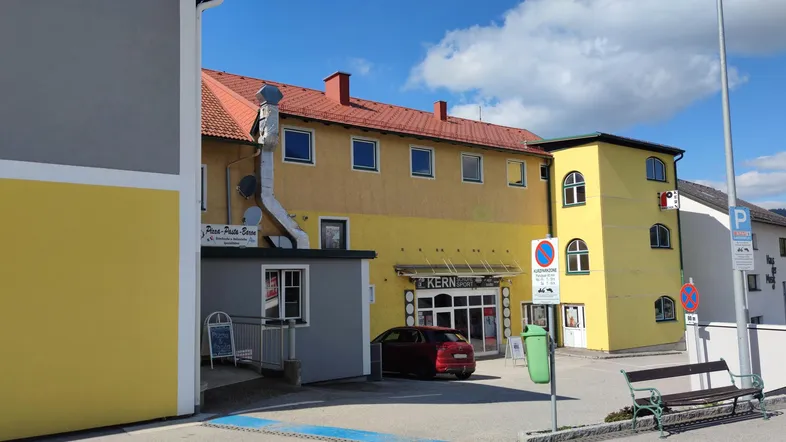 Gebäude mit gelber Fassade und rotem Dach, im Vordergrund ein kleiner Parkplatz und eine Bank.
